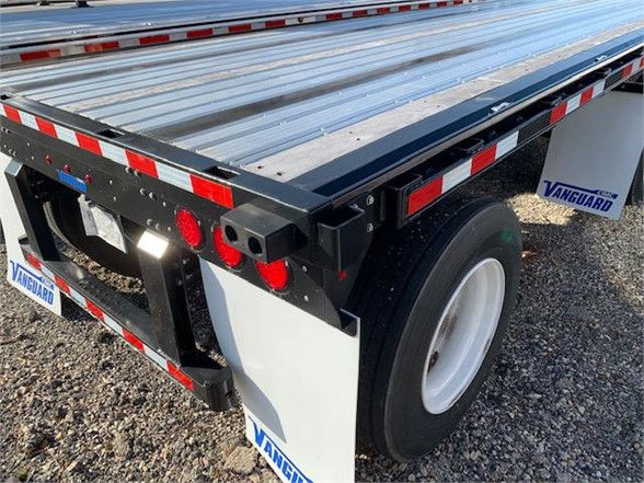 Vanguard 48ft steel flatbed trailer rear corner showing white wheel, black frame, red safety lights, reflective tape, and 'Vanguard' branding on mudflaps.