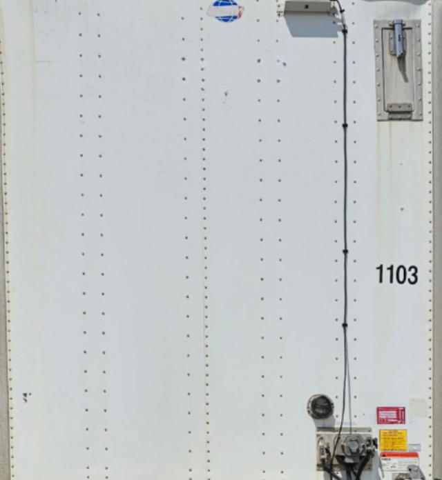 Trailer rear door area with riveted white exterior, number 1103, hydraulic connection and warning stickers, vertical cable along door.