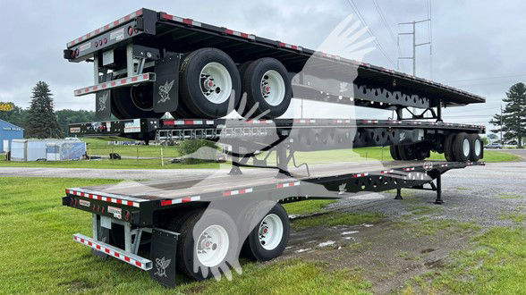 Manac steel flatbed trailer stacked on a split level with dual rear axles and white rims. Front of trailer shows reflective safety tape and multiple axles. VIN and stock number overlay in listing.