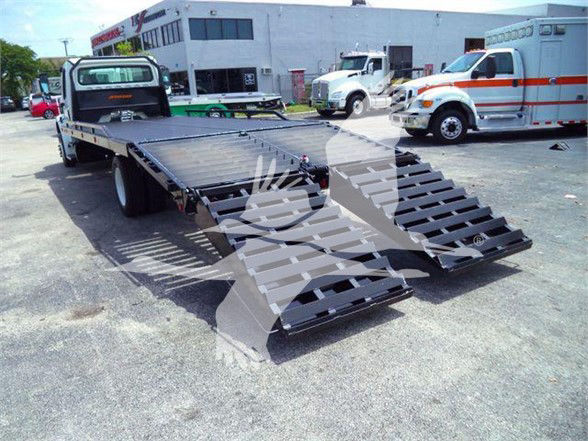 Flatbed tow truck with hydraulic ramps extended, Freightliner-style vehicle in background, equipment yard.