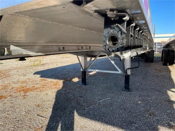 Flatbed trailer underside showing front landing legs and support frame with a metal I-beam structure and wheel well edge.