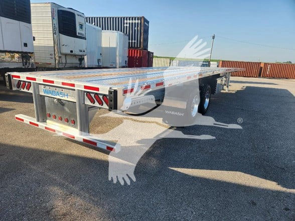Flatbed trailer by Wabash with reflective red/white tape, aluminum deck, dual rear lights, and spare tire on the side; stock lot with other trailers in background.