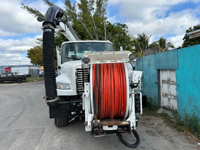 Front view of a white 2011 Freightliner M2 vacuum truck with a large red hose reel mounted on the front, black exhaust/hoses, and blue fence in background.