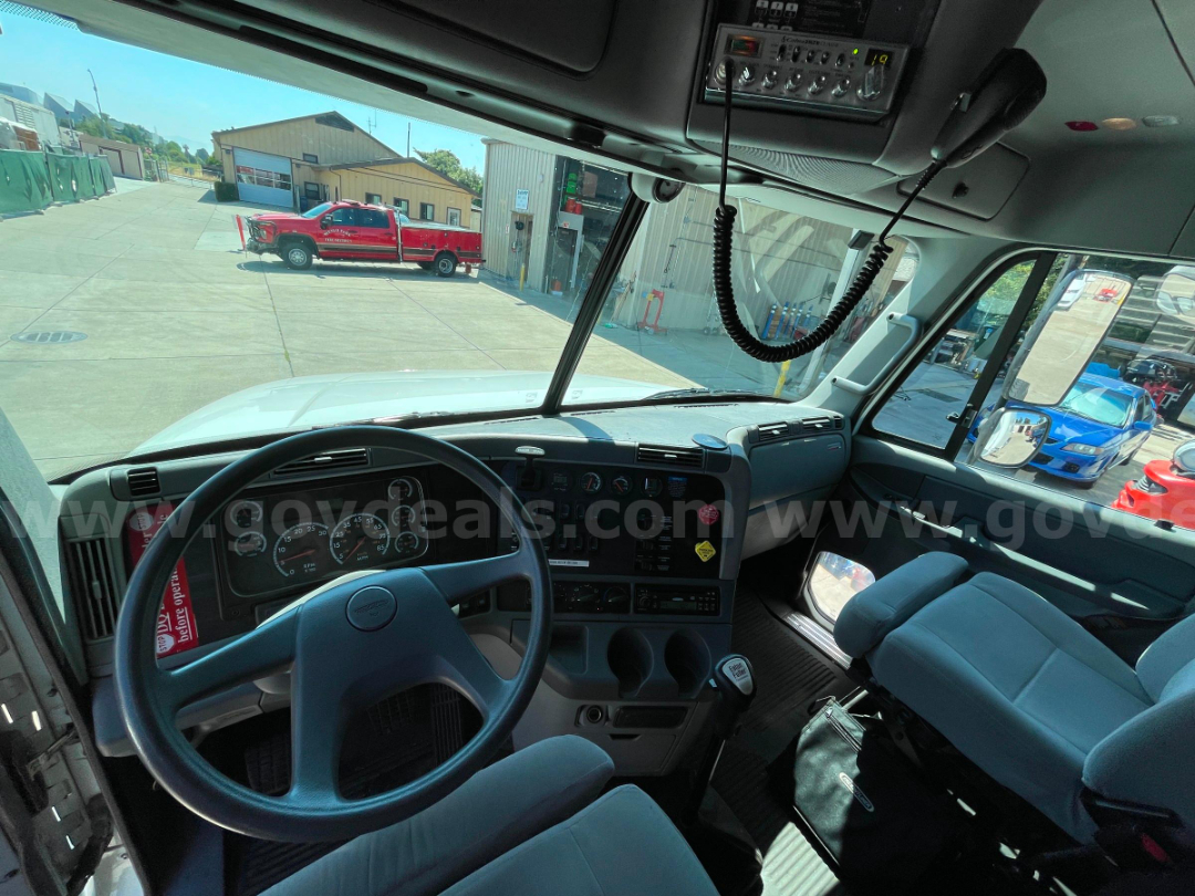 Truck cab interior showing steering wheel, dashboard gauges, central console, and driver seat. Window view of yard with trucks and buildings.