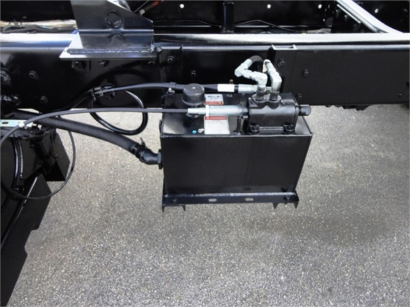 Truck chassis side with hydraulic pump and reservoir mounted under the frame, showing hoses and fittings.