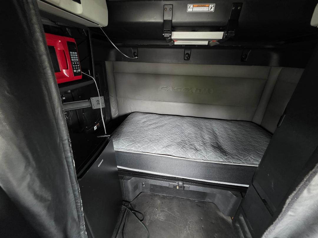 Truck cab sleeper area of a 2018 Freightliner Cascadia showing a single bed mattress, side storage, and a red microwave mounted inside the cab.