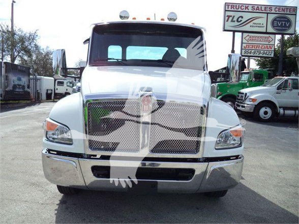 Kenworth T280 front view showing large grille, bumper, and side mirrors; dealership lot in background with trucks and signs.