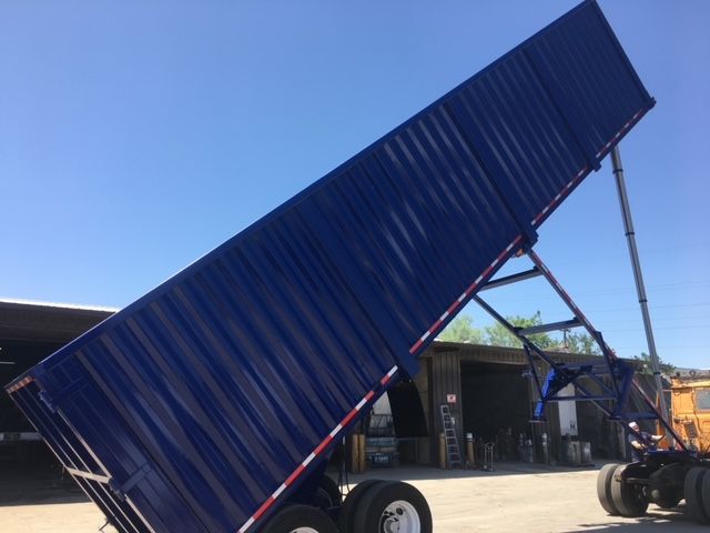 Trailer bed raised at a 45-degree angle showing blue rectangular dump trailer with dual rear axles and hydraulic lift in an industrial yard.