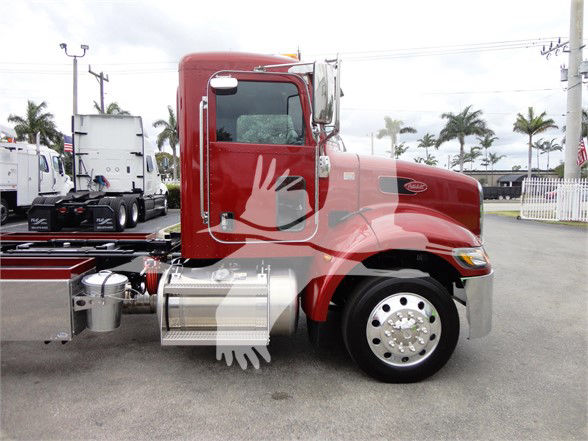 Side view of a red 2022 Peterbilt 337 tow truck with chrome fuel tanks and a large front wheel well, background show palm trees and other trucks.