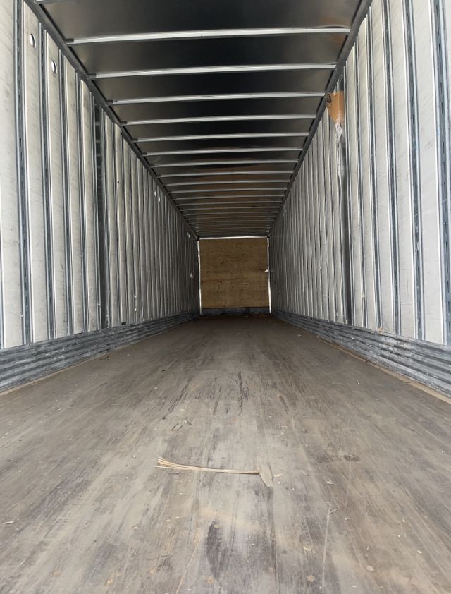Interior of a cargo trailer with metal walls and wooden floor, long and empty with a small piece of wood on the floor.