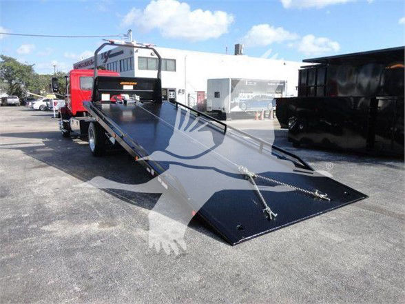 Tow truck with a flatbed tow platform extended, black metal bed, red cab, industrial yard background.