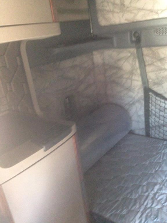 Cab interior of a 2012 Freightliner Cascadia sleeper with a bench-style seat and quilted bed. Built-in cabinetry and a small wardrobe area visible.