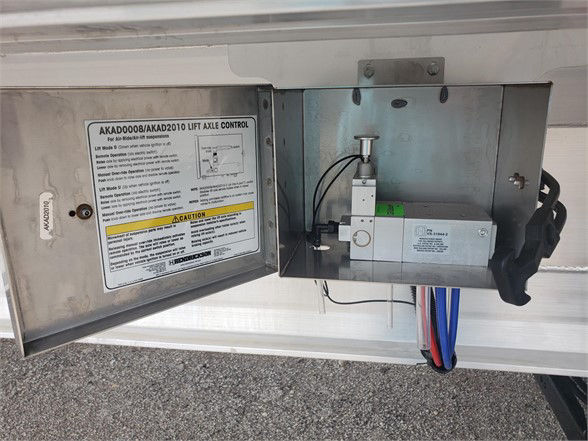 Flatbed trailer access panel showing AKAD000/AKAD2010 lift axle control label and a compact hydraulic/electrical control box with blue and red hoses.
