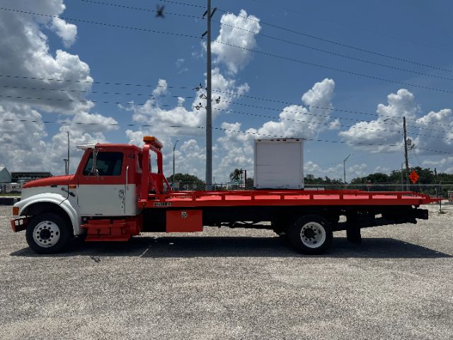 1999 International 4700 DT 466E tow truck with a long red flatbed and white cab, side view parked on gravel.