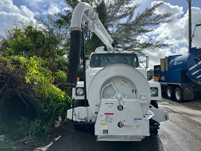 Front view of a white International vacuum truck with a large circular vacuum hose reel mounted at the rear and a hydraulic arm extending over the cab. License area and warning labels visible on reel housing.