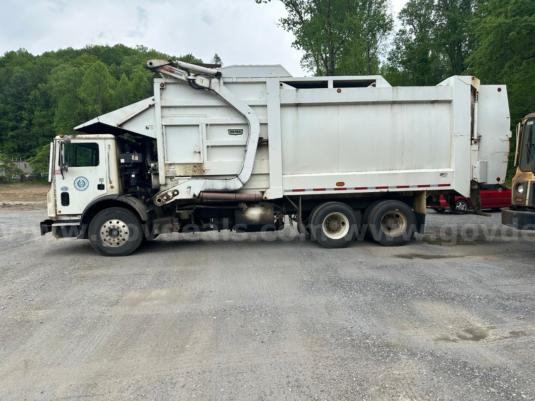 Side view of a 2017 Mack MRU613 garbage truck showing large rear dumpster and cab with hydraulic arms.
