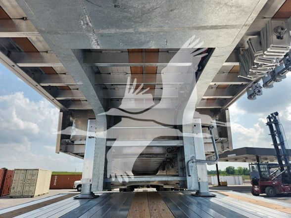 Under-trailer view showing the underside of a drop deck trailer with metal framework, hydraulic components, and rails.