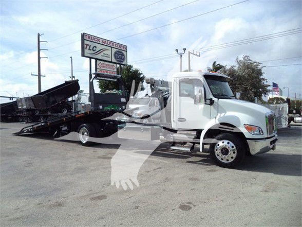 Tow truck with flatbed carrier and white cab, Kenworth truck in a lot.
