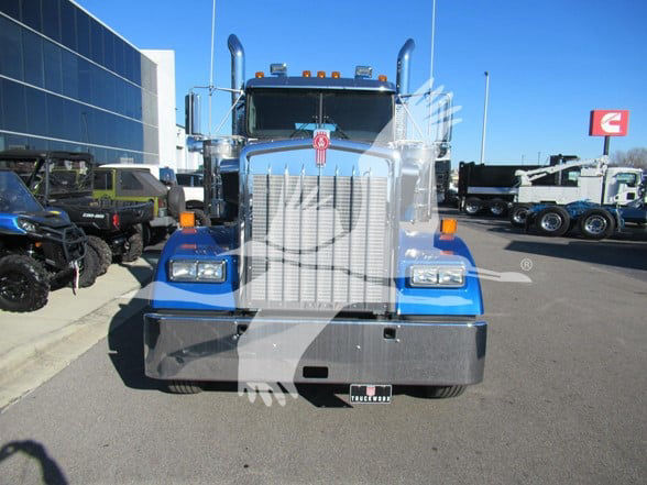 Kenworth W900L truck front view with prominent chrome grille, blue fenders, large bumper, and dual stacks. VIN plate, Kenworth emblem on hood, dealership lot in background.