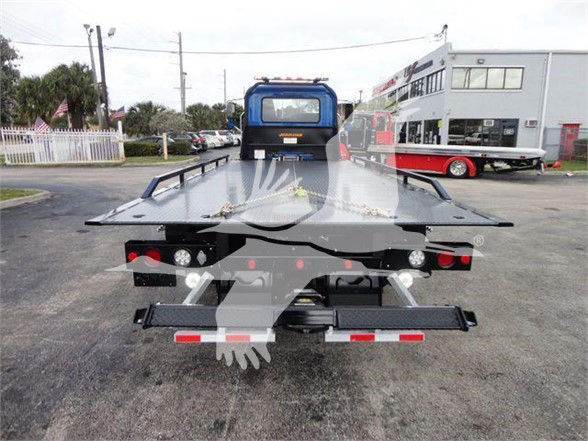 Tow truck flatbed rear view with blue cabin and black flatbed, lights on each side, license plate area, trailer hitch, and hydraulic mechanism visible.