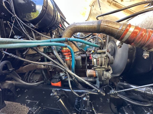 Engine bay with a turbocharger and belt-driven alternator, multiple blue/green hoses, red ignition coil, and thick orange-rimmed exhaust pipe in a heavy truck engine compartment.