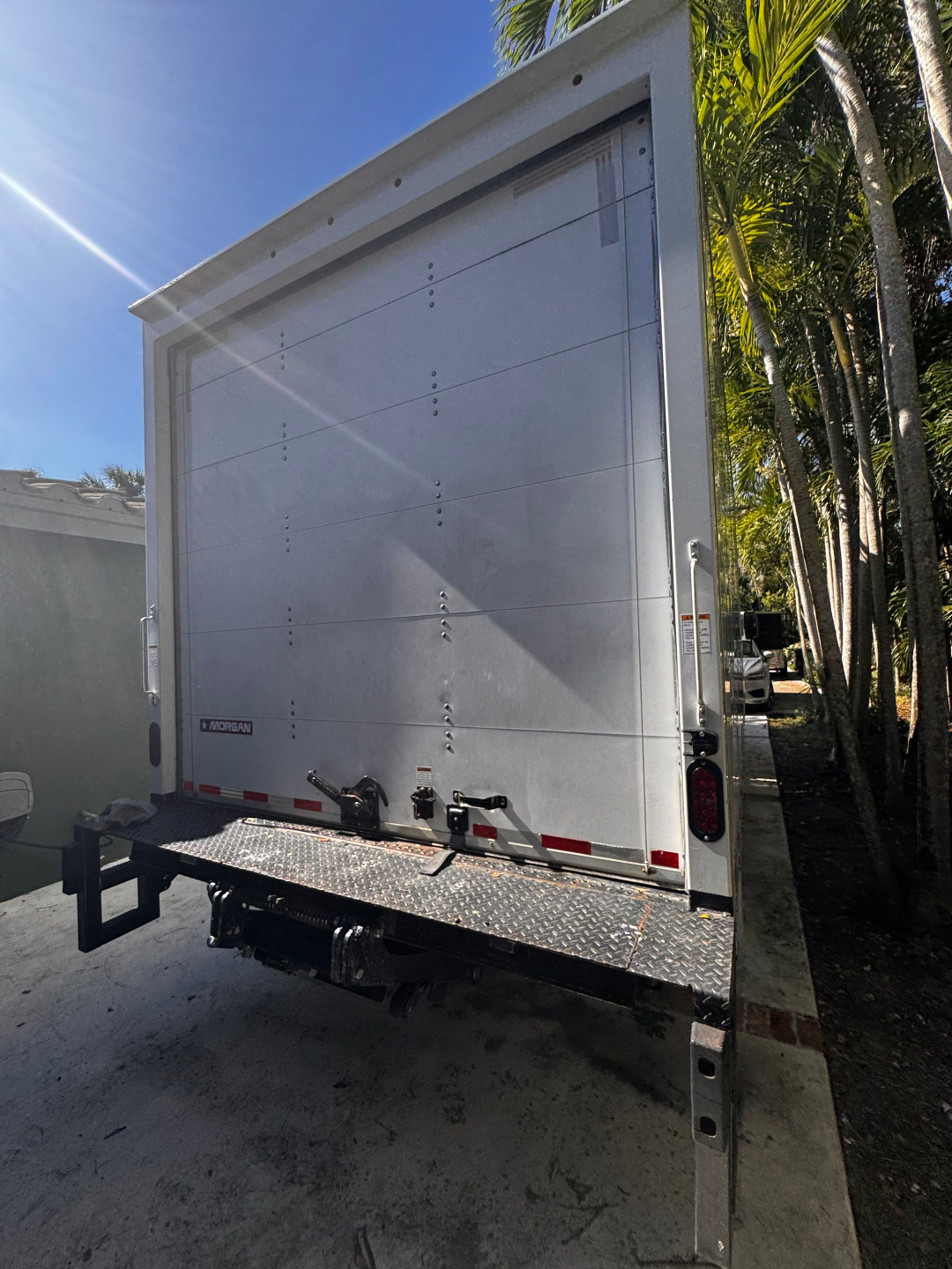 Rear view of a white Morgan Isuzu NQR box truck with a tall roll-up door, diamond plate bumper, and rear bumper with latches.