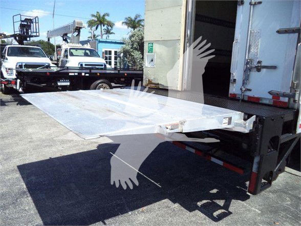 Flatbed truck ramp extending from a refrigerated trailer with metal surface and side support, dock area with other trucks in background.