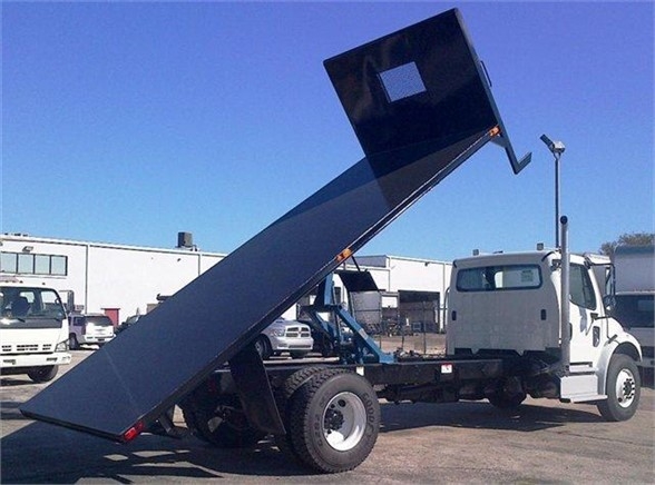 Flatbed truck body tilted at a 45-degree angle with a raised bed and rectangular cutout at the end, white cab, industrial lot in background.