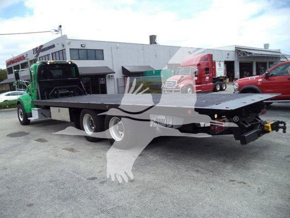 Flatbed tow truck on a lot with other trucks in background showing rear perspective and flatbed platform