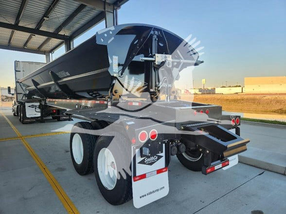 Smithco dump trailer rear three-quarter view showing hydraulic dump bed, dual axles with white wheels, taillights, and mud flaps. VIN sticker and stock number not visible on trailer.