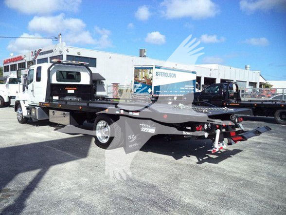 Tow truck with a flatbed, rear view showing loading ramps and winch. Some text on truck includes 'Ferguson' on a building in background and 'TOW' branding on equipment, unreadable small decals on flatbed.