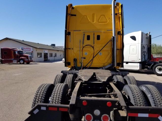 Rear view of a yellow Freightliner Cascadia truck cab on a trailer chassis, with black chassis and dual rear axles, other trucks in background.