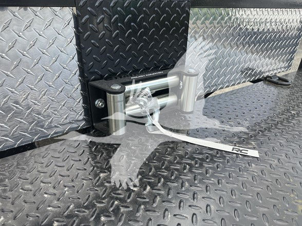 Trailer deck with diamond plate pattern and a chrome latch mechanism; shows a tether strap labeled RC attached near a silver hinge.