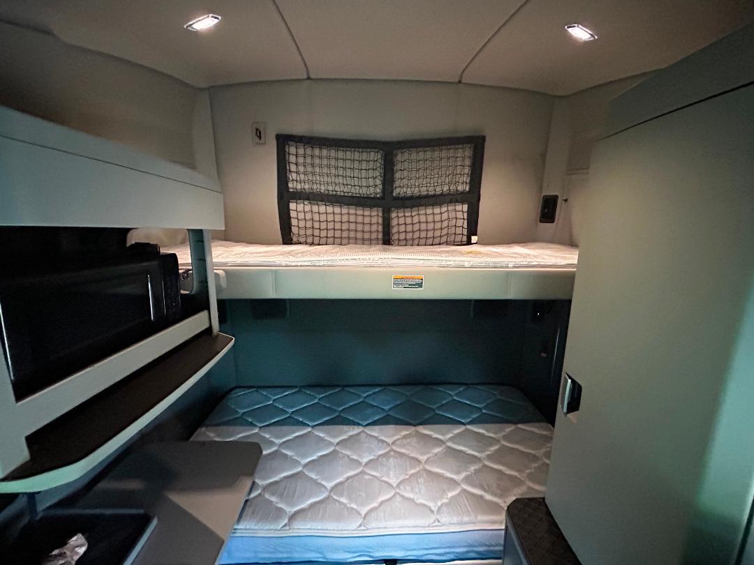 Truck sleeper bunk area in a 2020 Kenworth T680 showing upper bunk with net, lower bunk mattress, small desk area with microwave, and storage compartments.