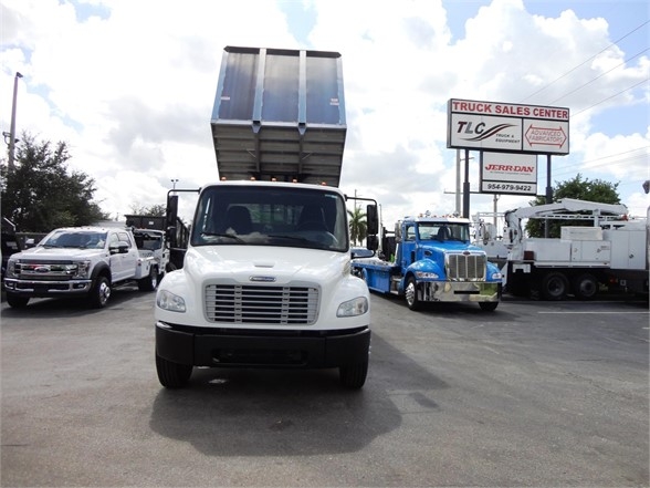 Front view of a white Freightliner Business Class M2 106 chassis with a raised dump bed, parking lot with other trucks in background.