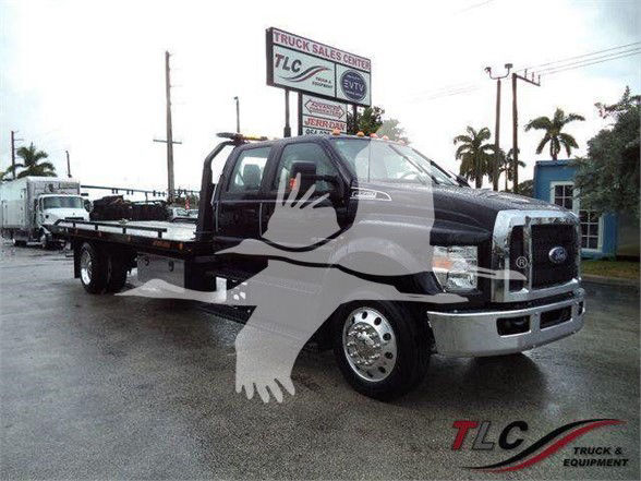 Ford F-750 tow truck flatbed showing cab with chrome grille and large side mirrors, TLC Truck & Equipment watermark, palm trees in background.