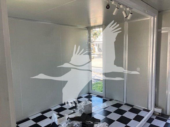 Interior trailer showroom with checkered black-and-white floor, white walls, track lighting, and large window. Decorative birds watermark overlays image.