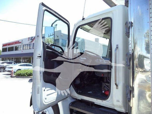 Truck cab interior with open door, steering wheel visible, seating area. Exterior shows part of a white cab and a tall chrome exhaust or equipment behind. No specific model text clearly legible in the image.