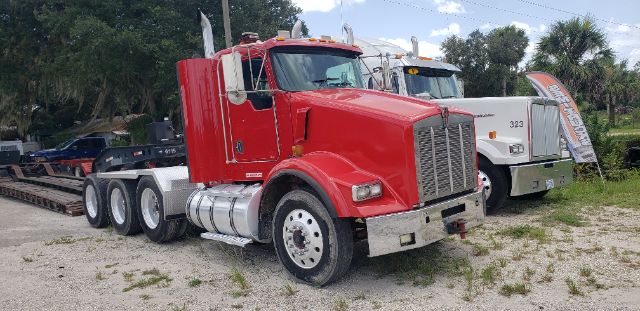 Kenworth tri-axle truck in red, front cab view, with chrome bumper and dual rear axles. Other truck in background.
