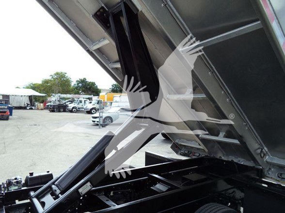 Dump truck bed raised showing hydraulic system and underside with metal supports and frame; some vehicles and trees visible in background.