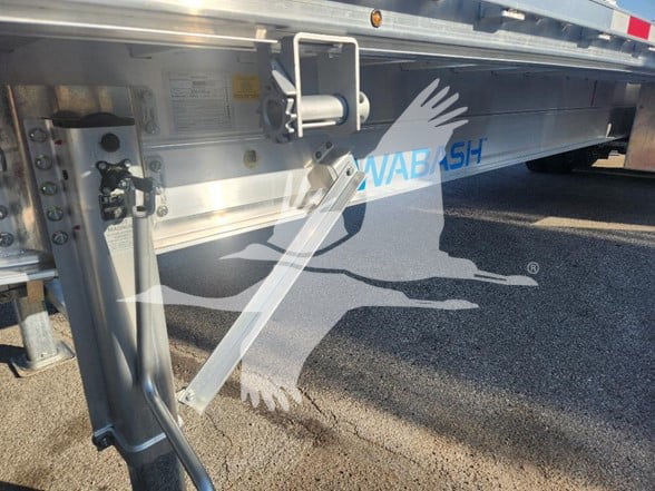 Flatbed trailer side showing WABASH logo in blue on corrugated aluminum side with support leg and ramp; hydraulic jack and stabilizer leg visible.