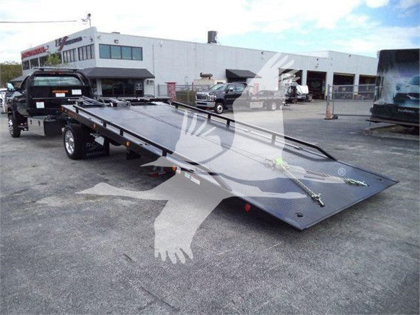 Tow truck trailer with flatbed, ramp folded down, showing V-shaped winch straps and hardware securing surface; background shows a white industrial building and other vehicles.