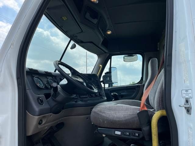 Truck cab interior of a 2015 Freightliner Cascadia showing steering wheel, dashboard, driver's seat with brown fabric, and door frame.