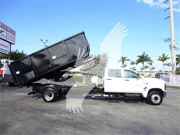 2023 Chevrolet Silverado 5500HD white crew cab with a black dump bed on a tilt trailer, parked in lot with palm trees in background.