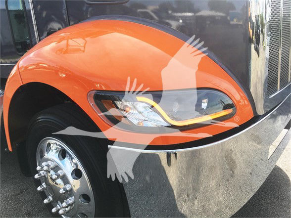 Front left fender and headlight area of an orange Peterbilt truck, wheel with chrome hub, part of the grille visible.