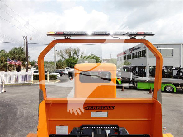 Rear view of an orange tow truck with a Jerr-Dan branded bed, overhead light bar, and reflection of a truck dealership lot.