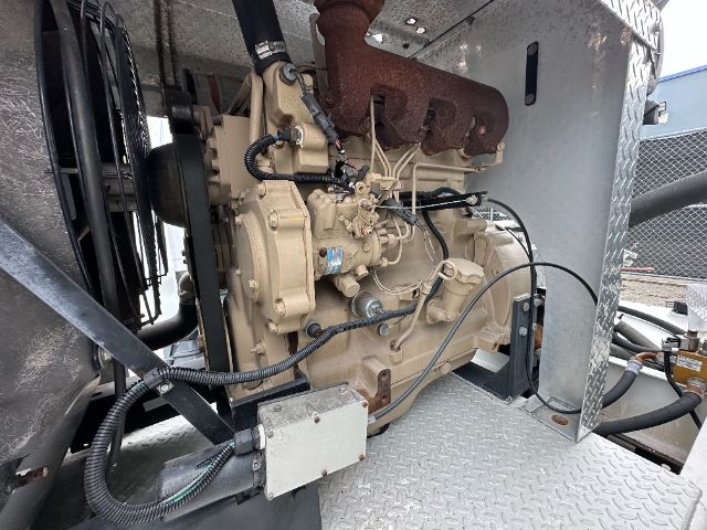 Engine bay of a GMC W5500 truck showing diesel engine with beige components, exhaust manifold, hoses, and wiring; mounted on a diamond-plate platform.
