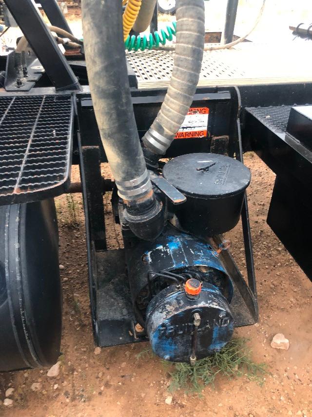 Tractor-trailer vacuum truck chassis with blue circular motor/pump, hoses, and caps; warning label visible on black tank, metal frame and tires in background.