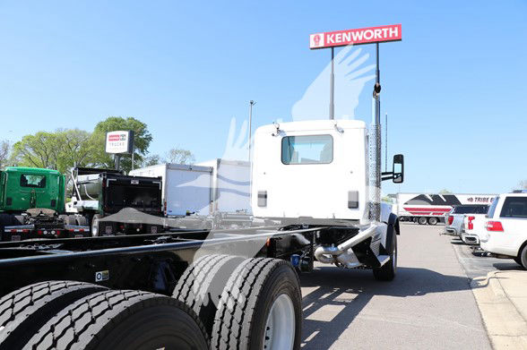 Kenworth T880 truck chassis with white cab, visible wheels, and Kenworth dealership sign in background.