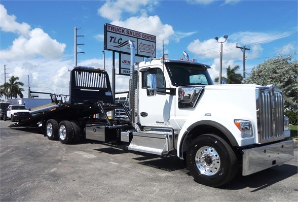 Kenworth W990 white tow truck with a heavy-duty flatbed and chrome accents, daytime outdoor dealership lot.